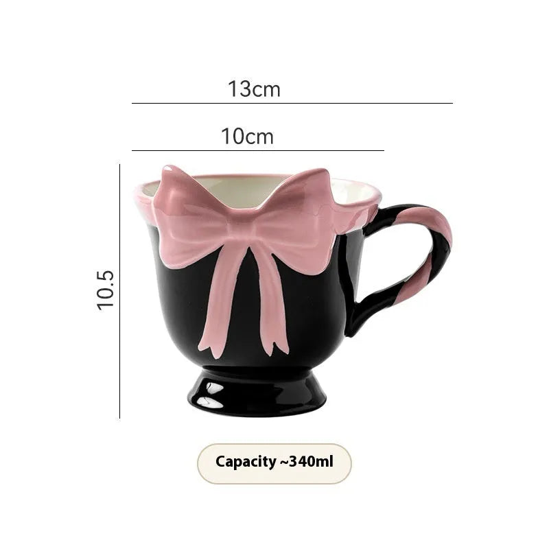 White ceramic mug with a heart and bow shaped handle, shown next to a black pink coloured version.