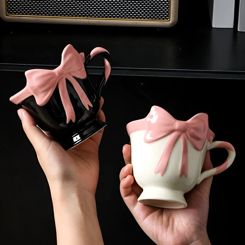 White ceramic mug with a heart and bow shaped handle, shown next to a black pink coloured version.