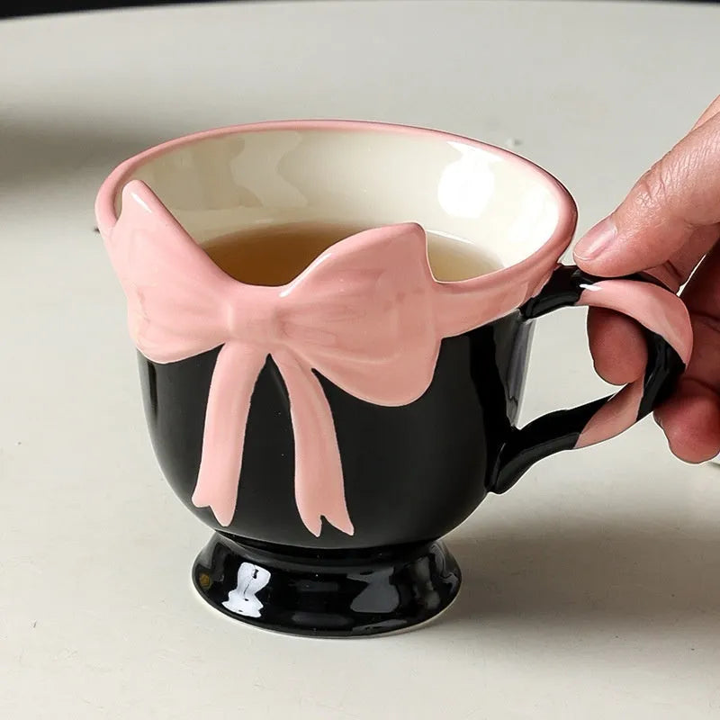 White ceramic mug with a heart and bow shaped handle, shown next to a black pink coloured version.