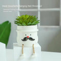 Small cartoon-style ceramic flowerpot with a raised hanging foot, on a desk next to a succulent.