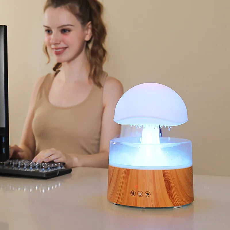 White cloud-shaped humidifier on a table emitting mist and showing coloured LED lights inside, with a remote control beside it.