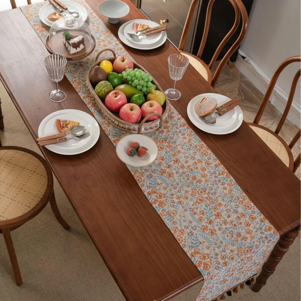 Beige jacquard table runner with long tassels and floral pattern laid on a wooden dining table.
