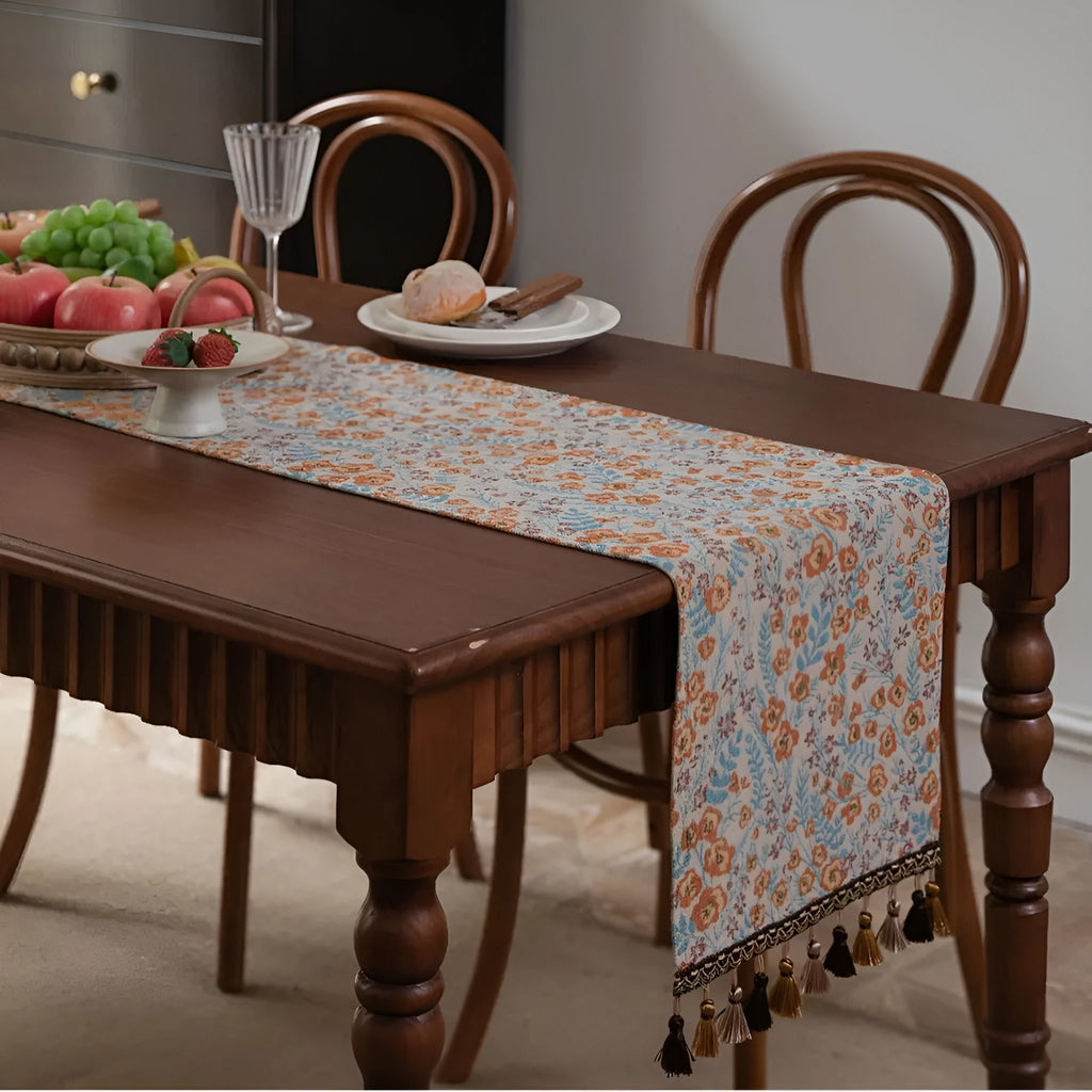 Beige jacquard table runner with long tassels and floral pattern laid on a wooden dining table.