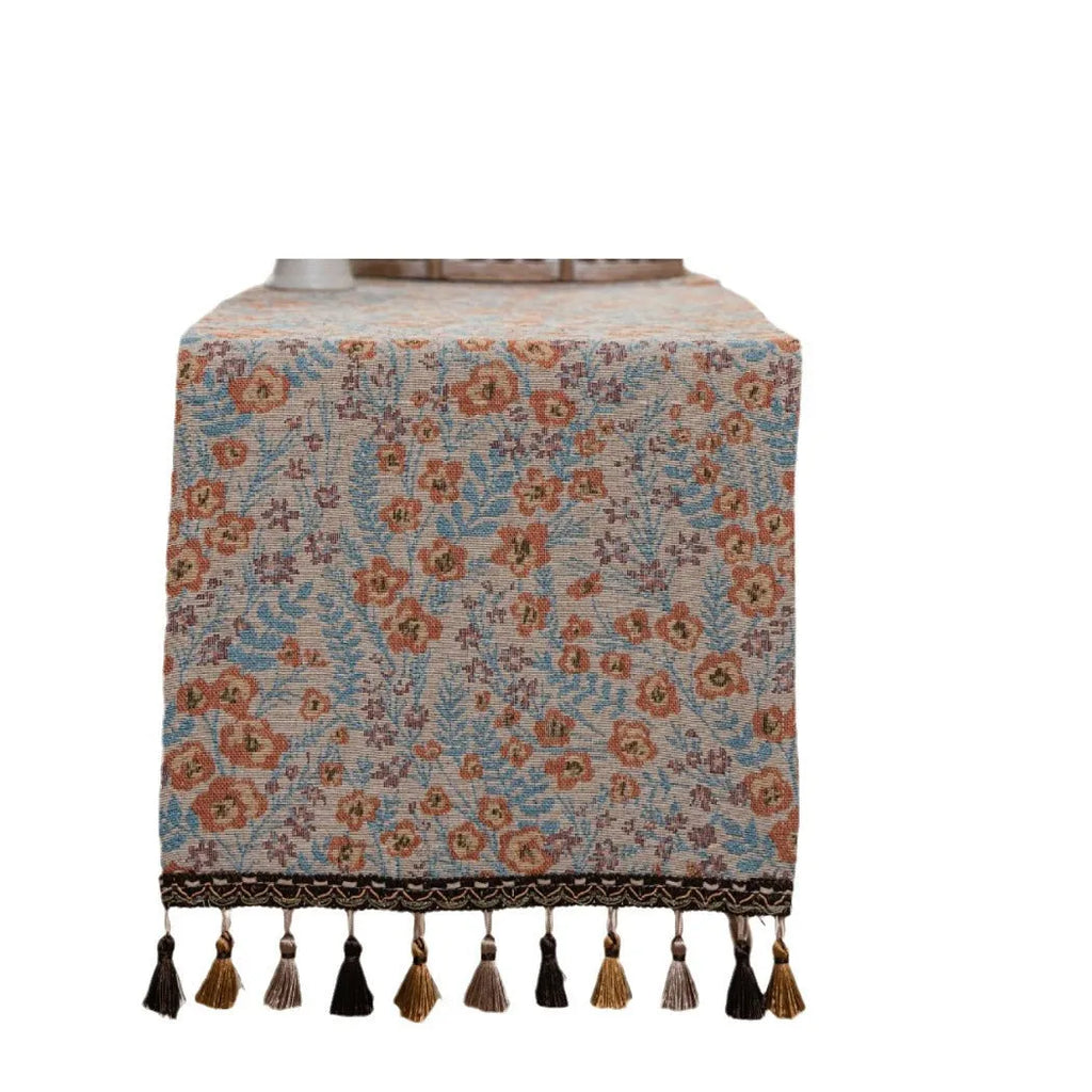 Beige jacquard table runner with long tassels and floral pattern laid on a wooden dining table.