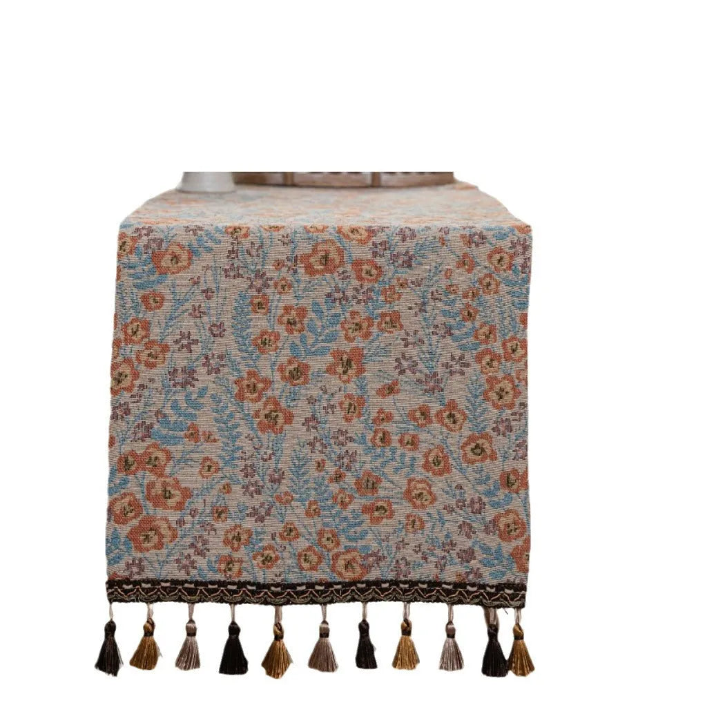 Beige jacquard table runner with long tassels and floral pattern laid on a wooden dining table.