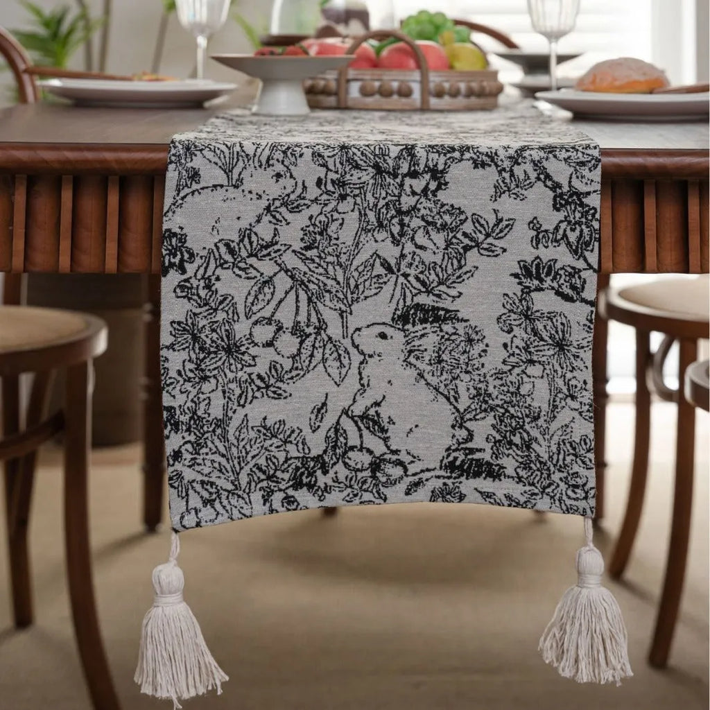 Beige jacquard table runner with long tassels and floral pattern laid on a wooden dining table.