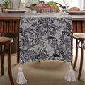 Beige jacquard table runner with long tassels and floral pattern laid on a wooden dining table.