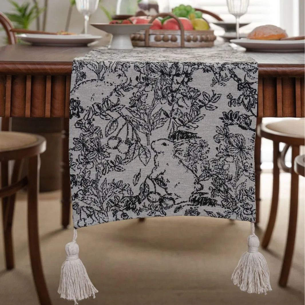 Beige jacquard table runner with long tassels and floral pattern laid on a wooden dining table.