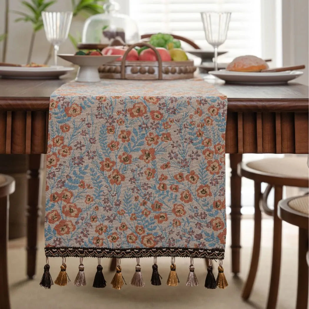 Beige jacquard table runner with long tassels and floral pattern laid on a wooden dining table.