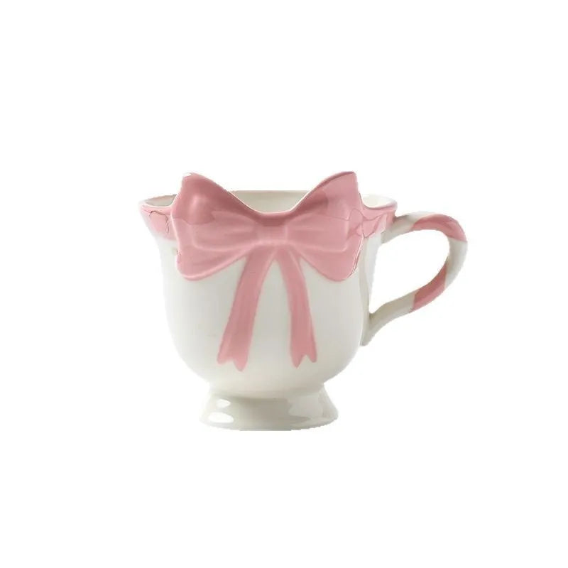 White ceramic mug with a heart and bow shaped handle, shown next to a black pink coloured version.