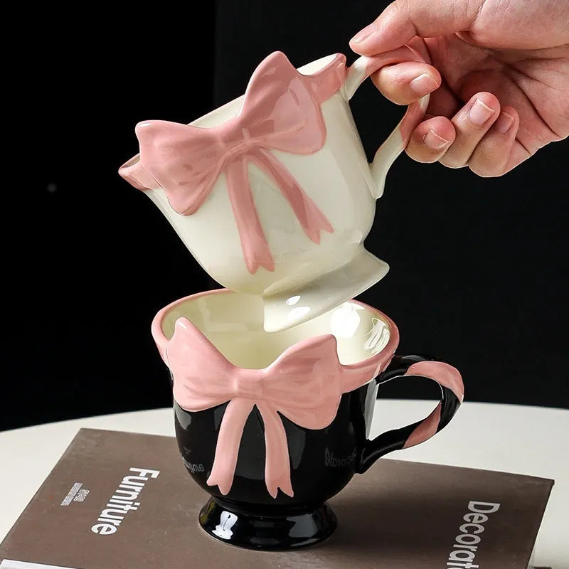 White ceramic mug with a heart and bow shaped handle, shown next to a black pink coloured version.