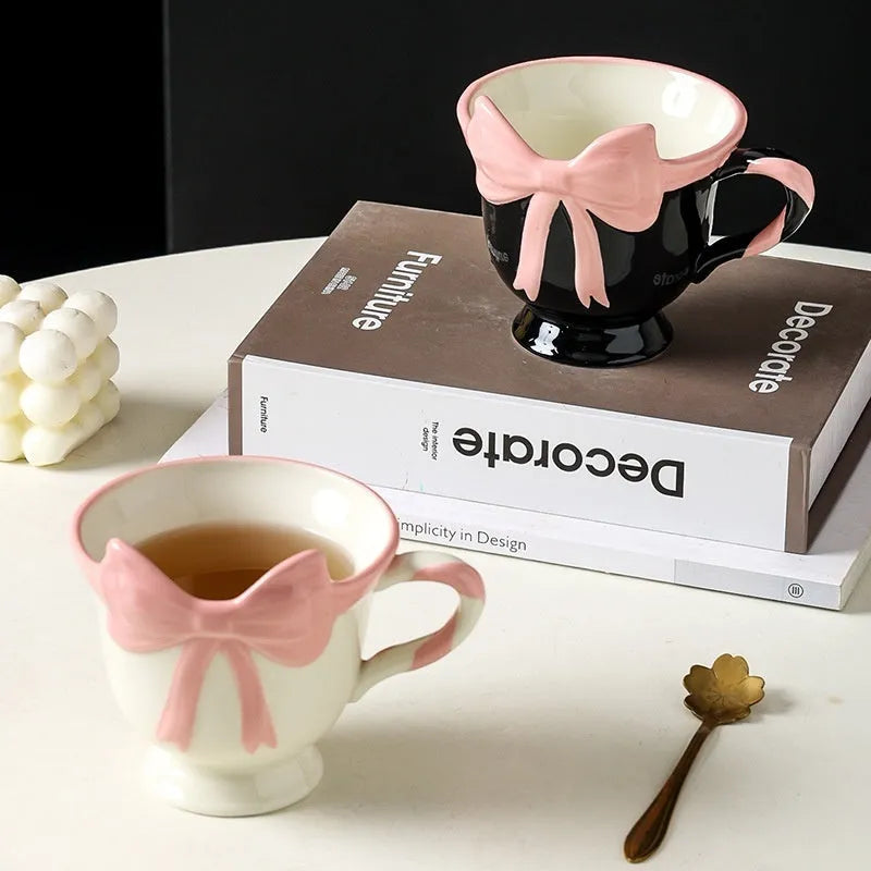White ceramic mug with a heart and bow shaped handle, shown next to a black pink coloured version.