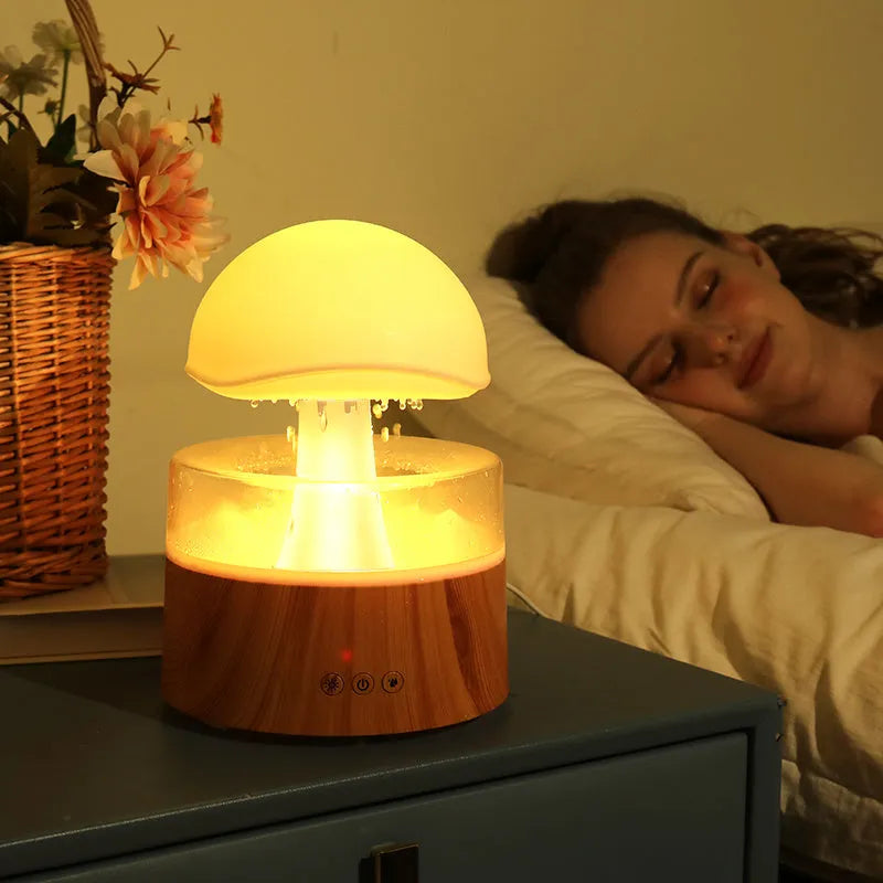 White cloud-shaped humidifier on a table emitting mist and showing coloured LED lights inside, with a remote control beside it.