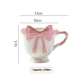 White ceramic mug with a heart and bow shaped handle, shown next to a black pink coloured version.