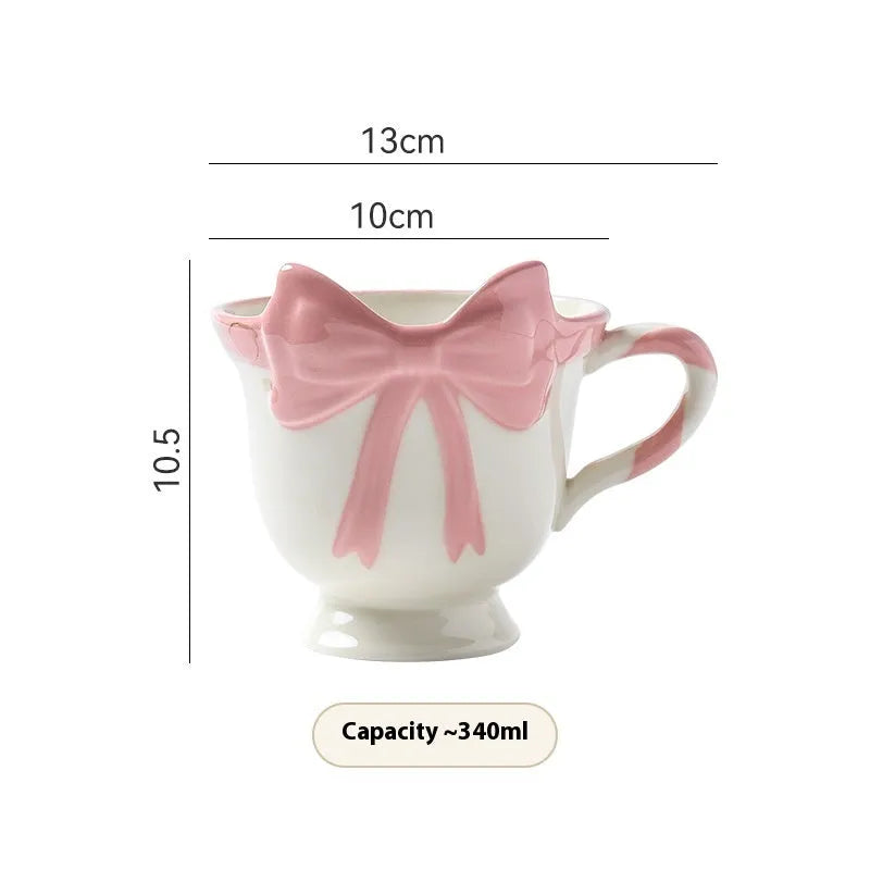 White ceramic mug with a heart and bow shaped handle, shown next to a black pink coloured version.